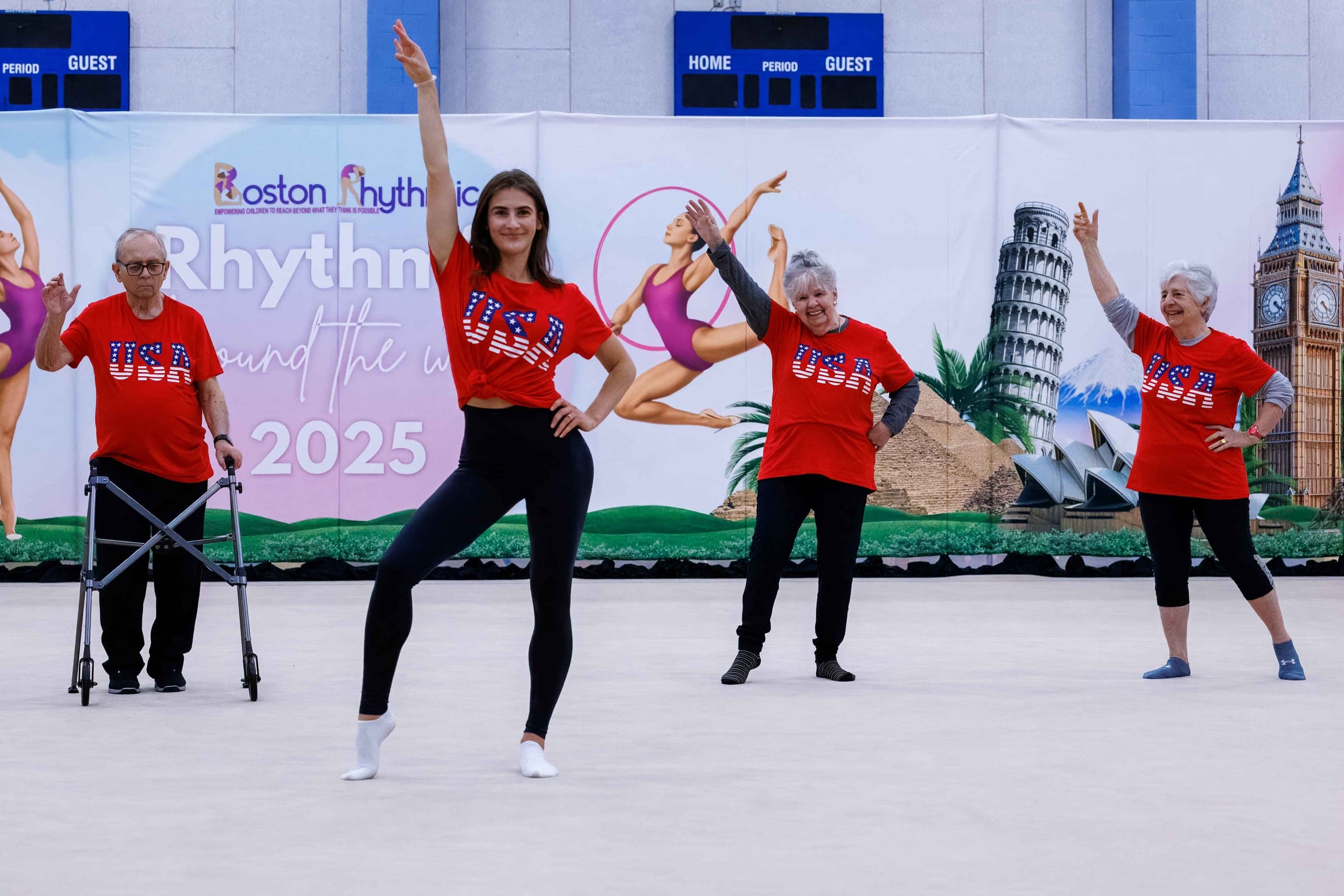 An instructor leading two seniors in red USA shirts through a dance routine in front of the Rhythmic Around the World backdrop.