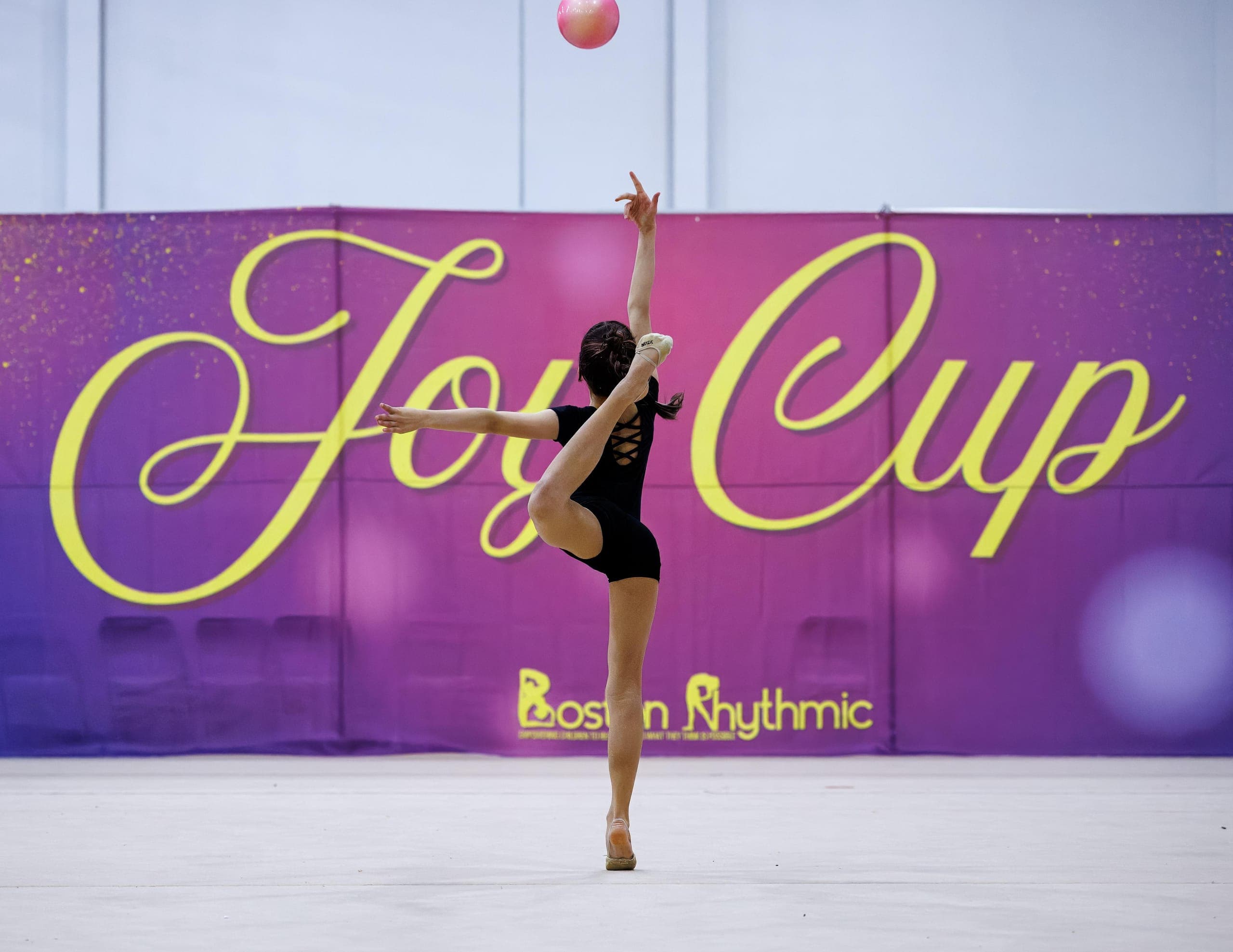 A Boston Rhythmic gymnast performing a ball routine in front of the purple and yellow Joy Cup event banner.