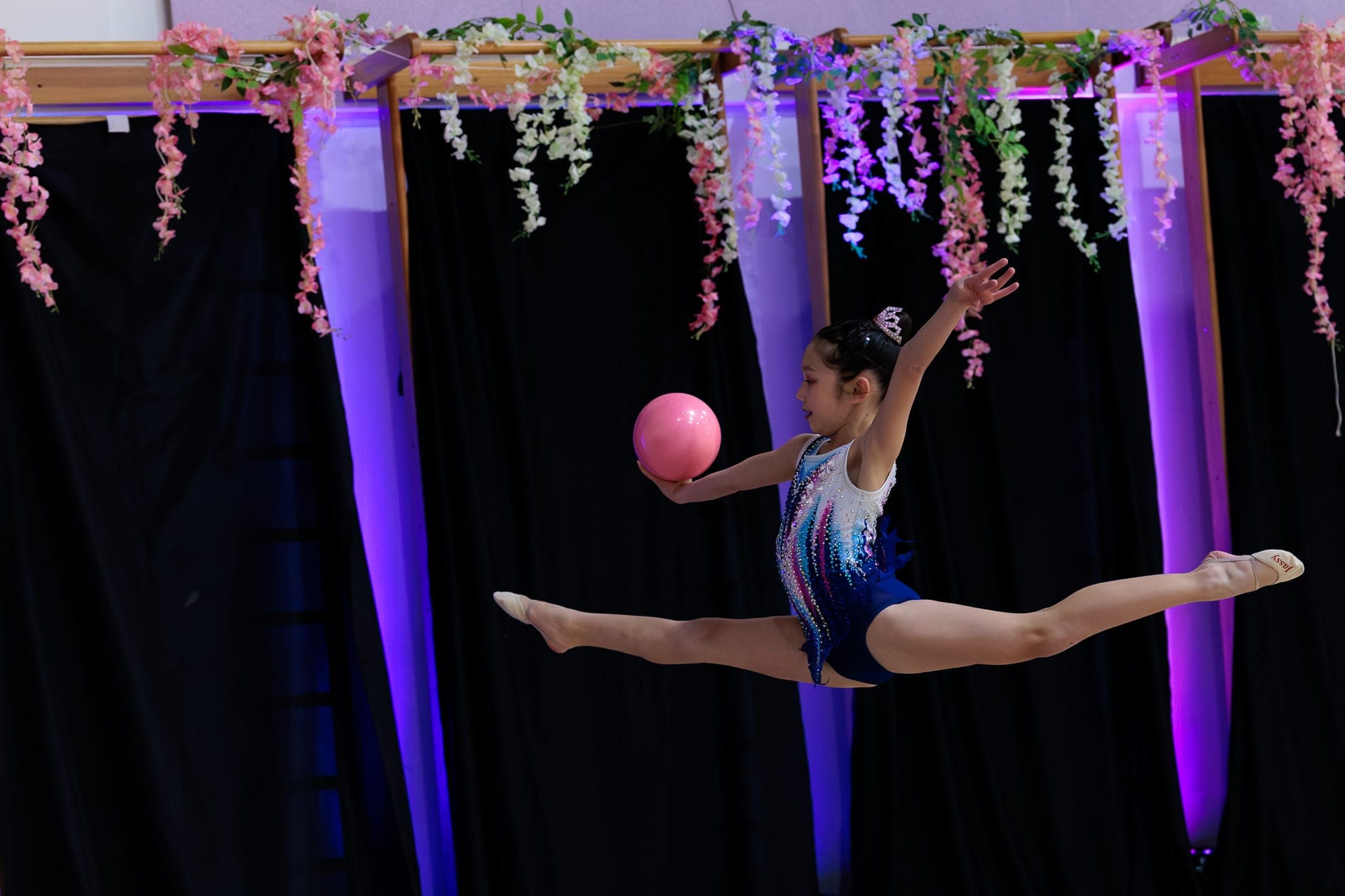 A Boston Rhythmic gymnast in a mid-air split leap holding a pink ball, framed by hanging floral decorations at the Blossom Cup.