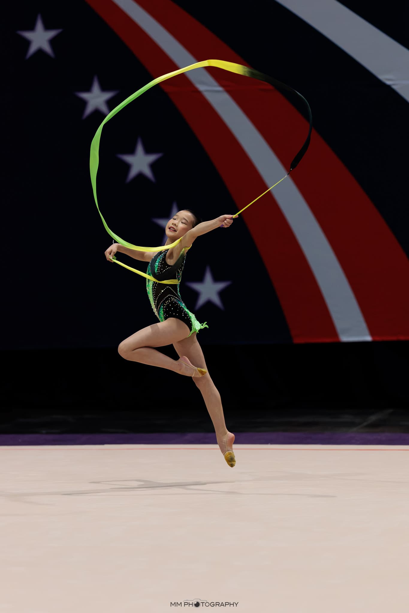 A gymnast mid-leap with a green and yellow ribbon at a national competition