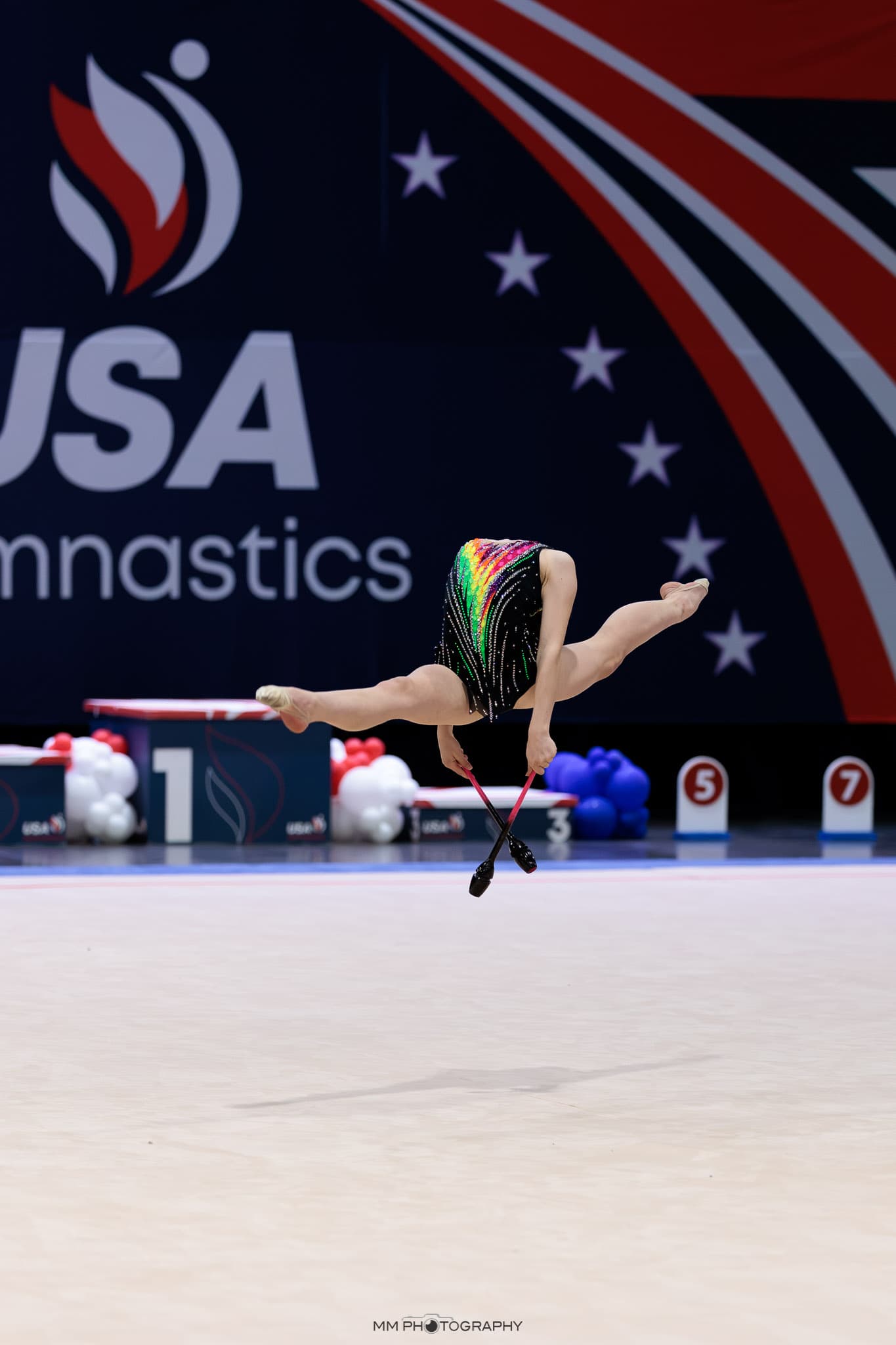 A gymnast in a mid-air backbend with clubs at a national competition