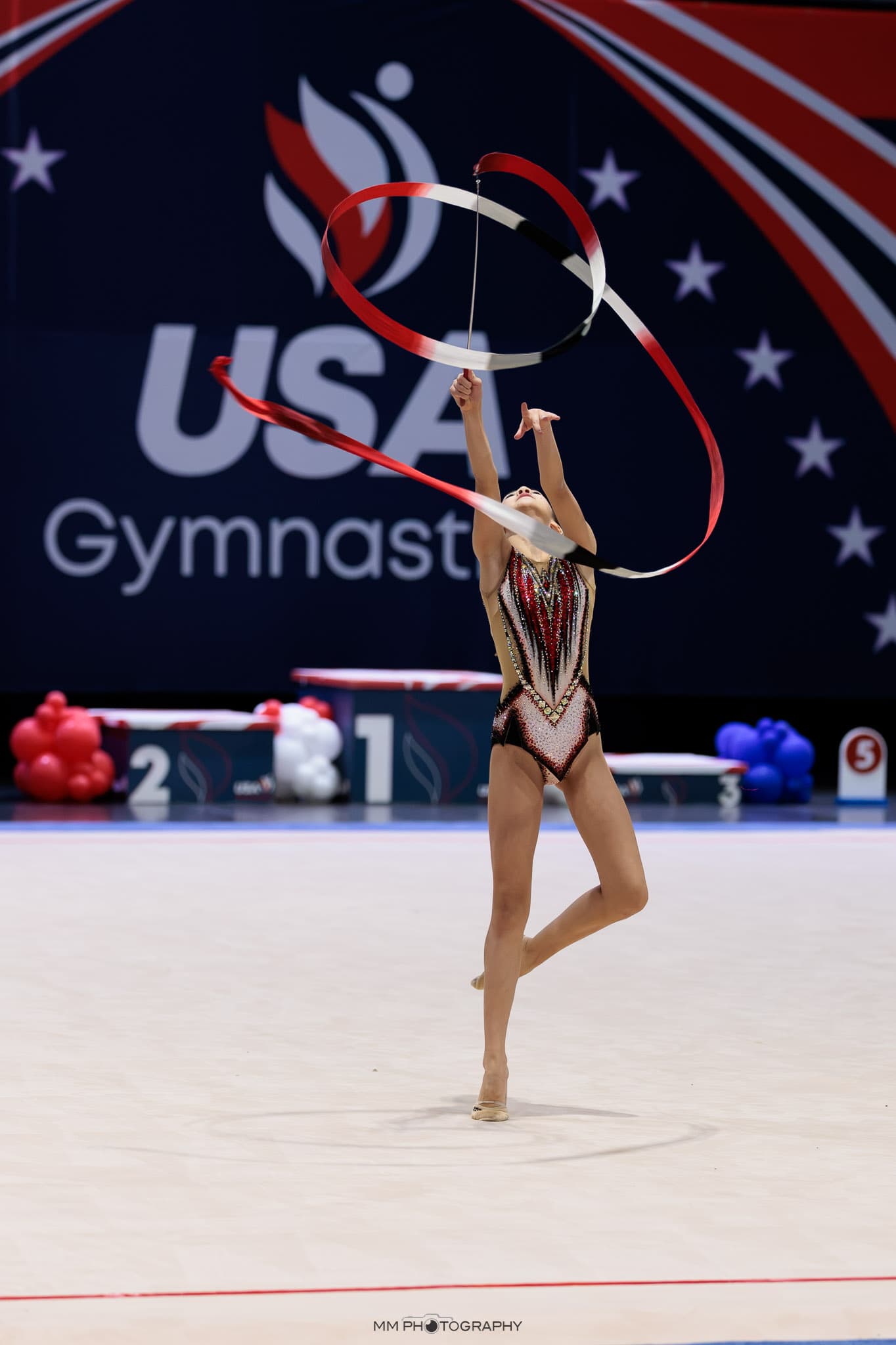 A gymnast reaching up as a red and white ribbon spirals overhead