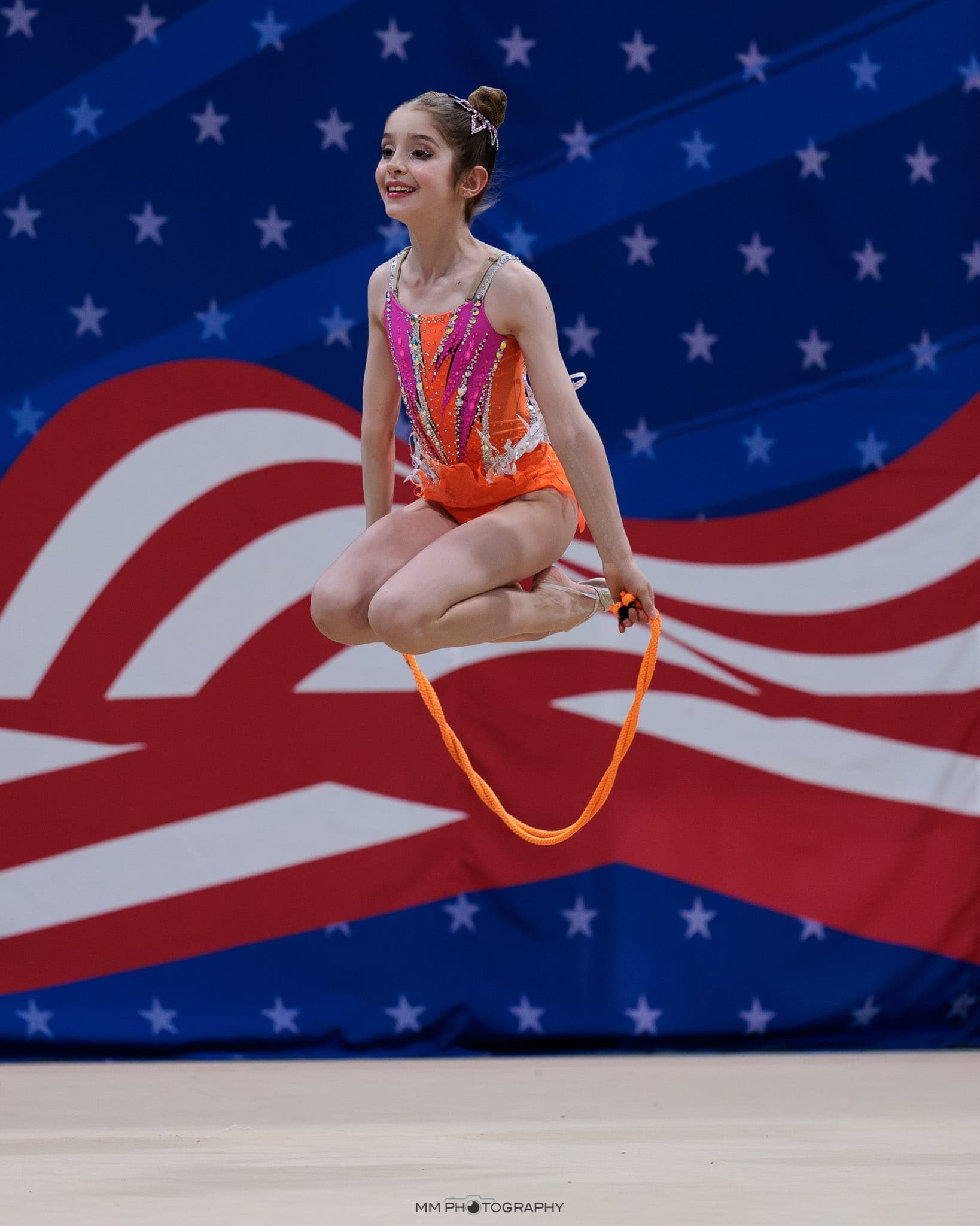A young gymnast in a tucked jump with an orange rope against an American flag backdrop